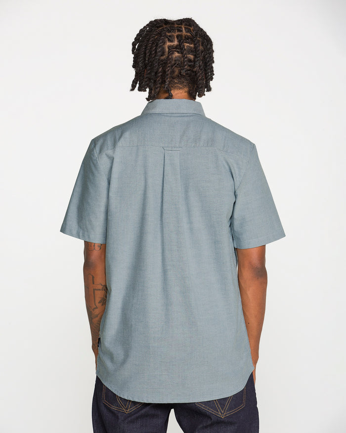 A person with short dreadlocks, wearing a light blue Volcom Men's Everett Oxford II Short Sleeve Shirt and dark jeans, is standing with their back to the camera against a plain white background.