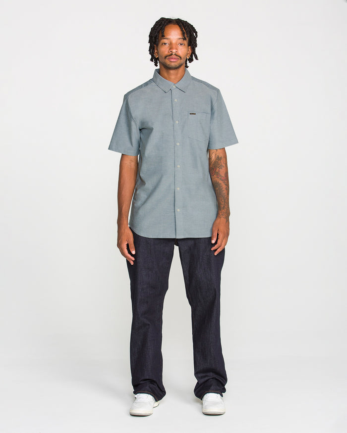 A man with short dreadlocks wears the Volcom Men's Everett Oxford II Short Sleeve Shirt in grey, styled with dark jeans and white sneakers, standing against a plain white background. This shirt’s stretch-woven fabric provides comfortable wear while maintaining a relaxed yet polished look.