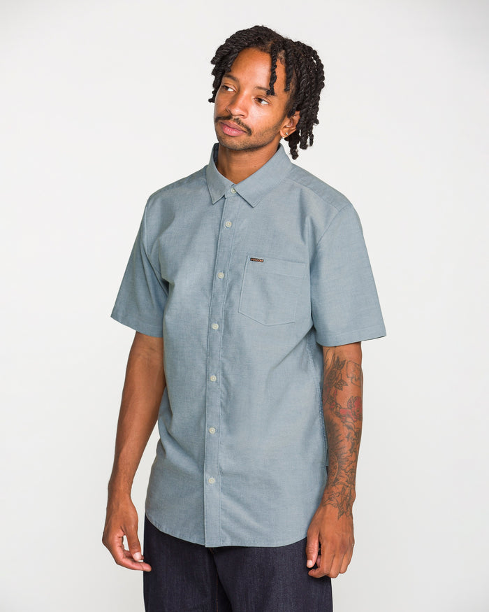 A man with short dreadlocks is wearing the Volcom Men's Everett Oxford II Short Sleeve Shirt in light blue, featuring a classic fit, chest pocket, and stretch-woven comfort for a relaxed modern vibe. He pairs it with dark pants while standing against a plain white background.