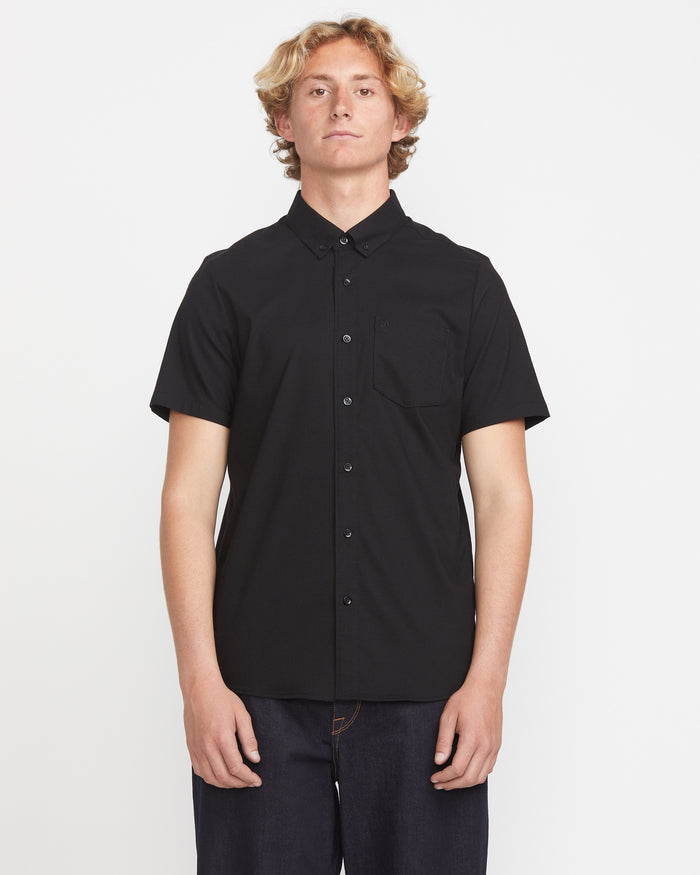 A young man with wavy blond hair wears the Volcom Men's Everett Oxford Short Sleeve, a black slim-fit button-up shirt with a chest pocket, standing against a plain white background.