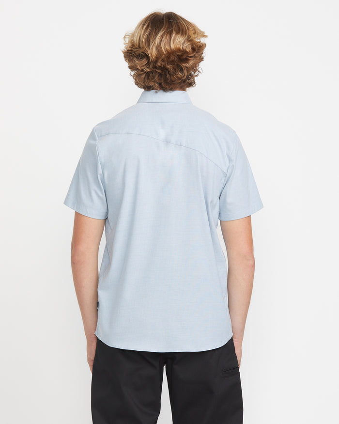 A person with wavy, light brown hair stands facing away from the camera, wearing the Volcom Men's Everett Oxford Short Sleeve in light blue and black pants against a plain white background.