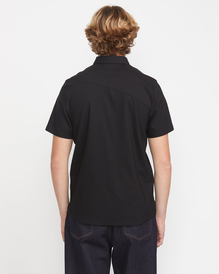A man with light brown, wavy hair faces away from the camera, wearing the Volcom Men's Everett Oxford Short Sleeve in black and dark jeans, standing against a plain white background.