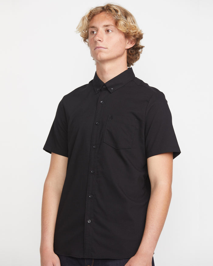 A young person with wavy blond hair wears the Volcom Men's Everett Oxford Short Sleeve, a slim-fit black shirt with a pocket, standing against a plain white background and looking slightly to the side.