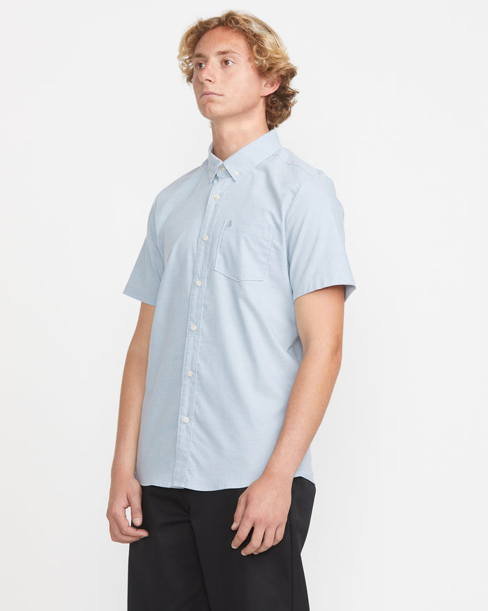 A man with wavy blonde hair wears the Volcom Men's Everett Oxford Short Sleeve in light blue and black pants, standing against a plain white background and looking slightly to the side.