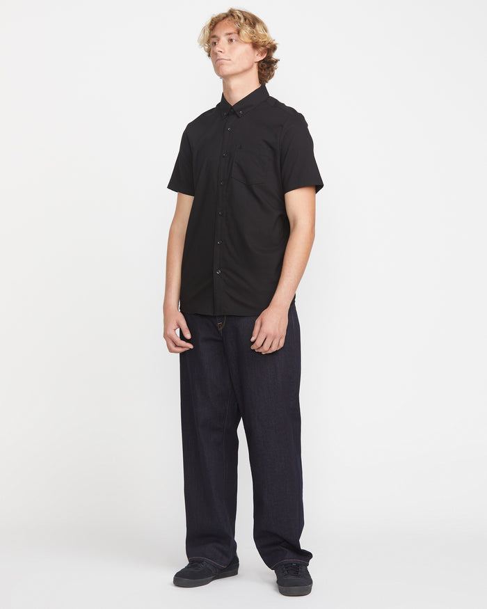 A young man with light, wavy hair and a neutral expression wears the Volcom Men's Everett Oxford Short Sleeve in black, paired with loose dark pants and black shoes, standing against a plain white background.