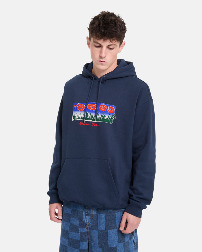 A young person wearing a navy blue Volcom Men’s Featured Artist Antonio Carrau Pullover hoodie with a colorful graphic, paired with patterned blue jeans, stands against a plain white background.