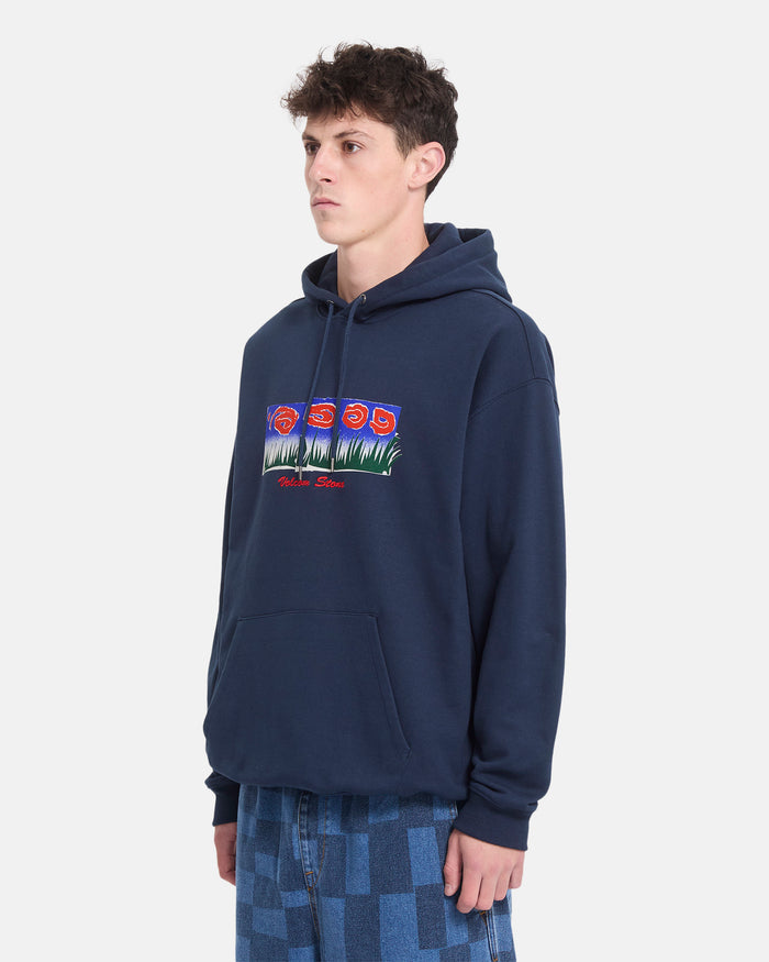 A young man stands against a white background, looking to the side, wearing Volcom’s Men’s Featured Artist Antonio Carrau Pullover in navy with a floral graphic, paired with blue checkered pants.