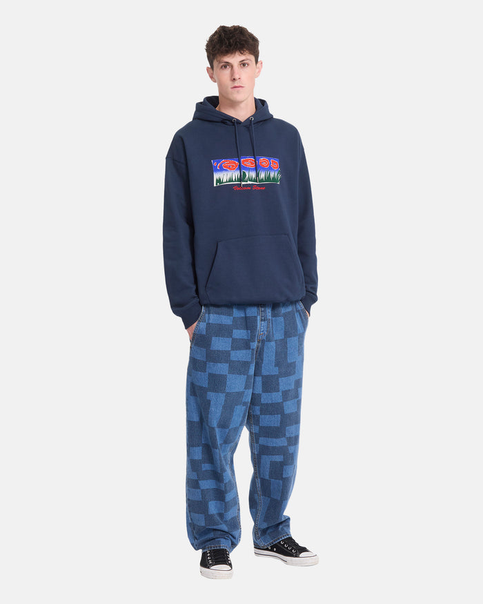 A young man wearing a Volcom Men’s Featured Artist Antonio Carrau Pullover in navy, paired with heavyweight fleece, blue checkered baggy jeans, and black sneakers stands facing forward against a plain white background.