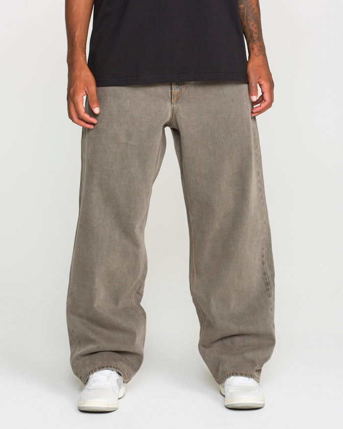 A person stands against a plain white background, cropped at the shoulders, wearing Volcom Men's Featured Artist Antonio Carrau Reverb Pants in gray, paired with a black overdyed cotton T-shirt and white sneakers.