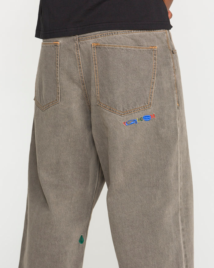 A person is seen wearing the Volcom Men's Featured Artist Antonio Carrau Reverb Pants—loose, gray-toned jeans with visible orange stitching, a vibrant embroidered motif near the right back pocket, and a small green accent by the left hem. Their upper body is partly shown in a black shirt.
