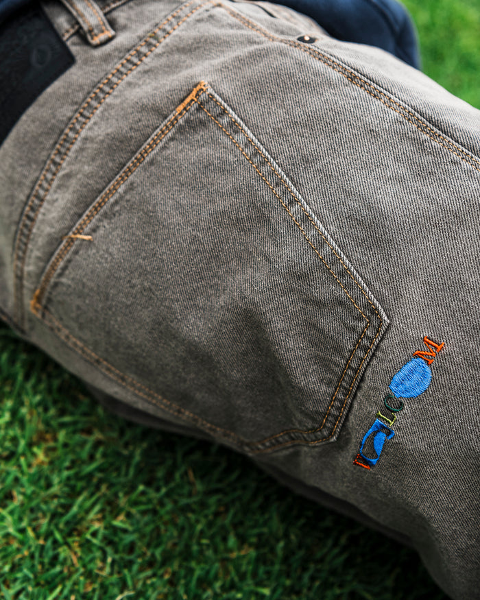 A close-up of the Volcom Men’s Featured Artist Antonio Carrau Reverb Pants in gray denim showcases orange stitching and colorful embroidery near the pocket, as part of a person lies on green grass. The low-rise fit highlights their casual style, while subtle black cotton detailing adds a modern touch.