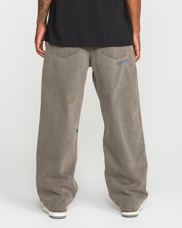 A person is standing with their back to the camera, wearing Volcom Men’s Featured Artist Antonio Carrau Reverb Pants—loose-fitting, low-rise gray jeans with small embroidered details on the back pockets—and a black shirt made of overdyed cotton. White shoes are partially visible.