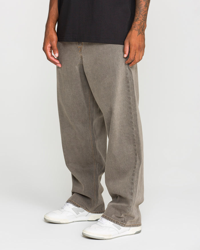 A person is shown from the waist down, standing against a plain white background and wearing Volcom Men's Featured Artist Antonio Carrau Reverb Pants in overdyed black cotton. The loose-fitting, low-rise silhouette of the pants is highlighted, paired with a black shirt and white sneakers.