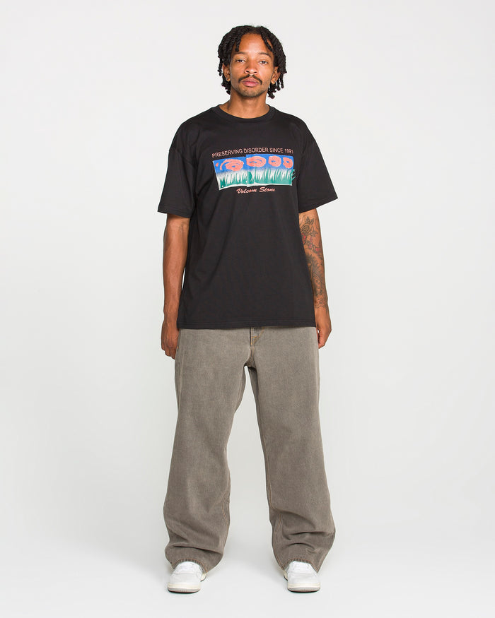 A man with short dreadlocks stands against a plain white background wearing a black graphic T-shirt, white sneakers, and Volcom Men's Featured Artist Antonio Carrau Reverb Pants in loose gray overdyed black cotton.
