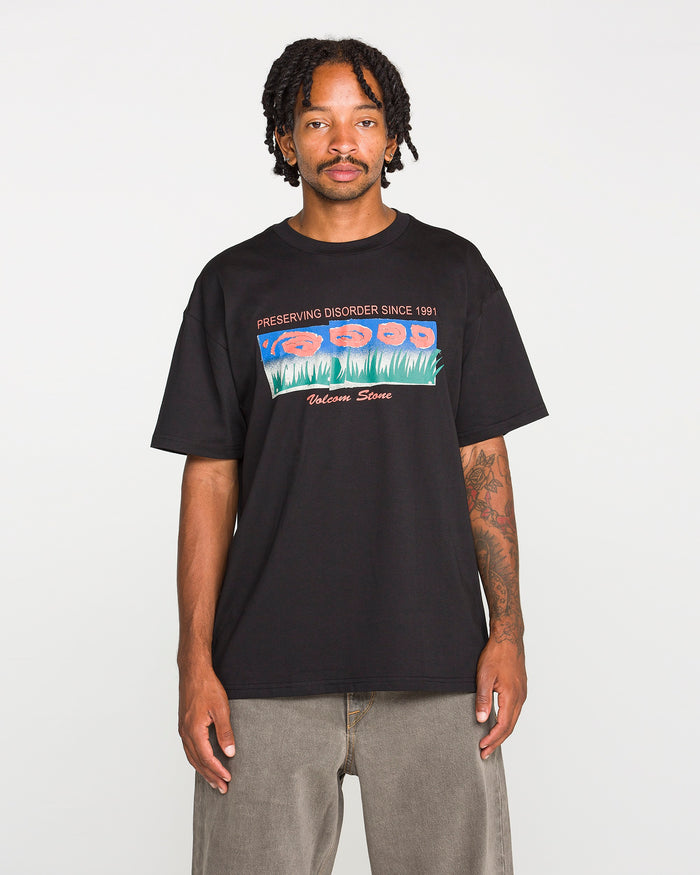 A man stands facing forward against a plain white background, wearing the Volcom Men's Featured Artist Antonio Carrau Short Sleeve Tee—a premium black shirt adorned with colorful graphics and the text "PRESERVING DISORDER SINCE 1991" and "Welcome Stone.