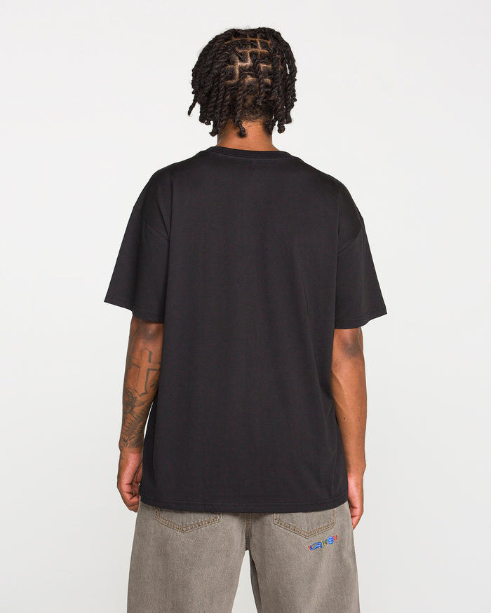 A person with short dreadlocks is standing with their back to the camera against a plain white background, wearing the Volcom Men's Featured Artist Antonio Carrau Short Sleeve Tee in black and gray jeans.