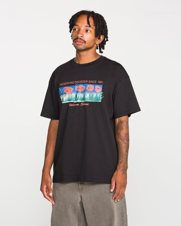 A person wearing the Volcom Men's Featured Artist Antonio Carrau Short Sleeve Tee, a premium black shirt showcasing colorful collage artwork graphics and the text "PRESERVING DISORDER SINCE 1991" and "Welcome Sons," stands against a plain background.
