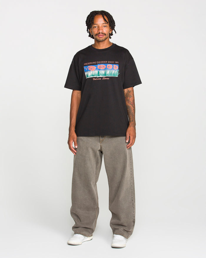 A man with short dreadlocks wears the Volcom Men's Featured Artist Antonio Carrau Short Sleeve Tee—a premium black t-shirt showcasing a bold graphic design—paired with baggy gray jeans and white sneakers, standing facing forward against a plain white background.