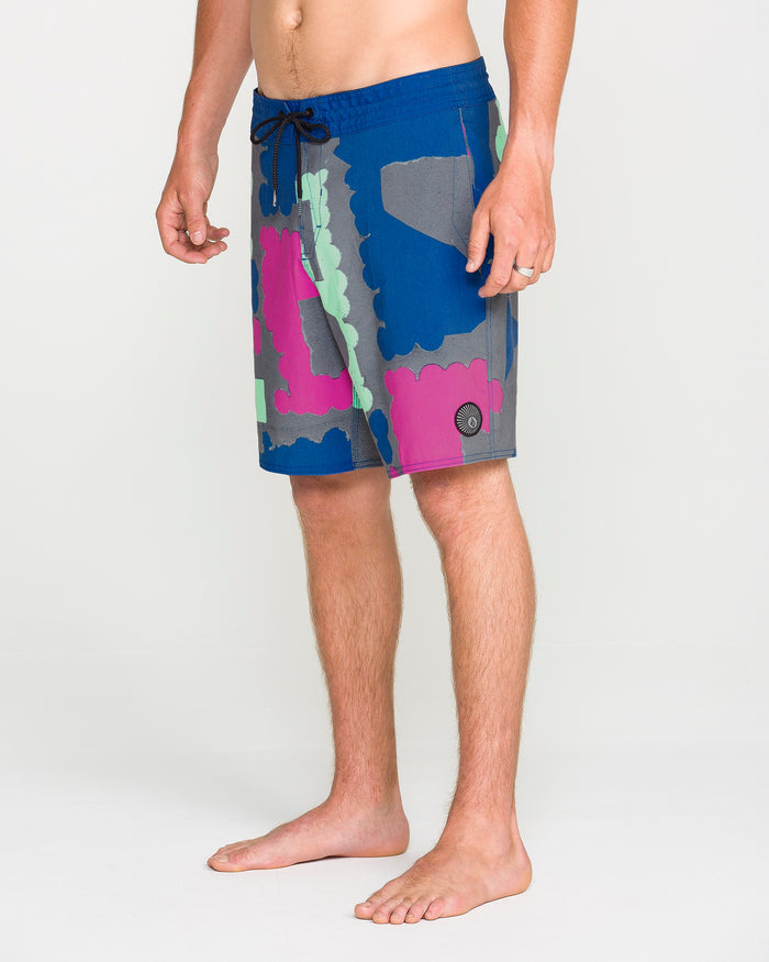 A man stands barefoot, wearing Volcom Men's Featured Artist Antonio Carrau Stoney Trunks—colorful knee-length board shorts with a blue, pink, green, and gray abstract pattern. The shorts feature a black Cinch Fly drawstring and a small circular Volcom logo near the hem.