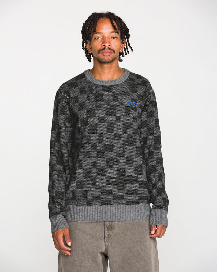 A person with short dreadlocks wears the Volcom Men's Featured Artist Antonio Carrau Sweater—black and gray jacquard—in addition to loose gray pants, standing against a plain white background and facing the camera with a neutral expression.