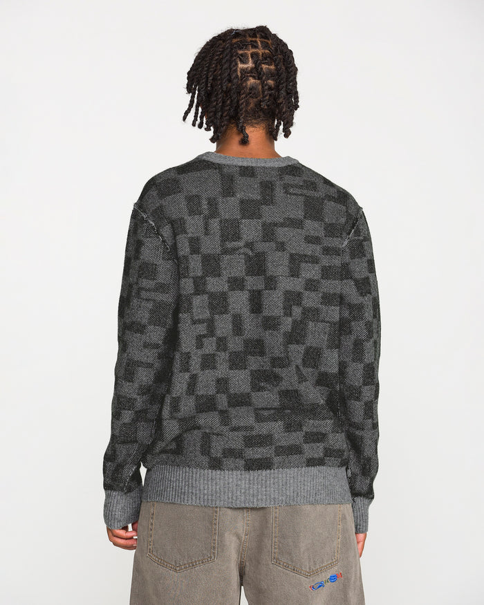 A person with short, twisted hair is seen from behind against a plain white background, wearing the Volcom Men's Featured Artist Antonio Carrau Sweater in dark gray checkered jacquard paired with brown pants.