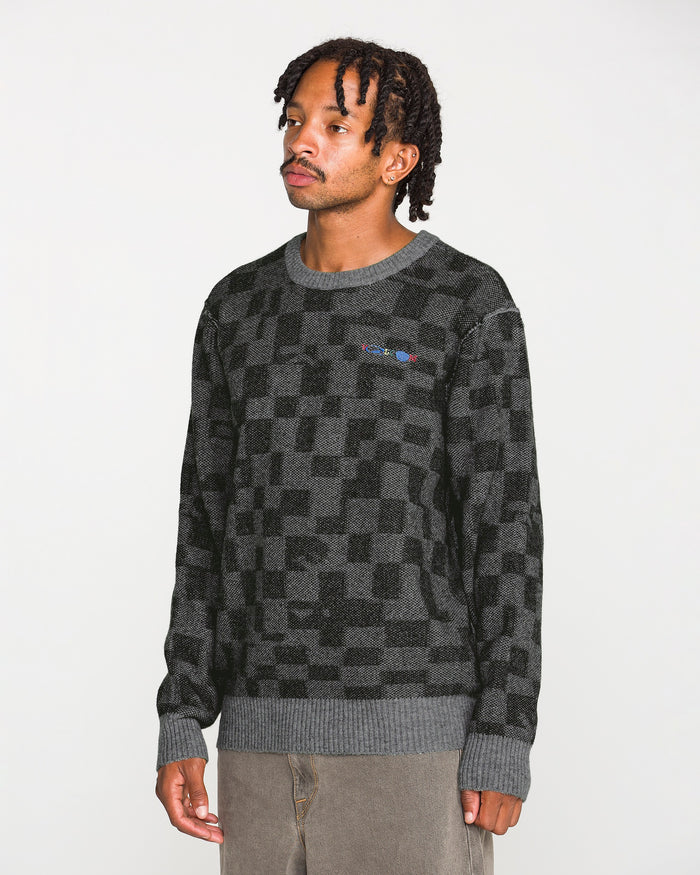 A man with short dreadlocks is wearing the Volcom Men's Featured Artist Antonio Carrau Sweater—a dark gray jacquard piece with ribbed cuffs and hem—paired with light brown pants, standing against a plain white background.