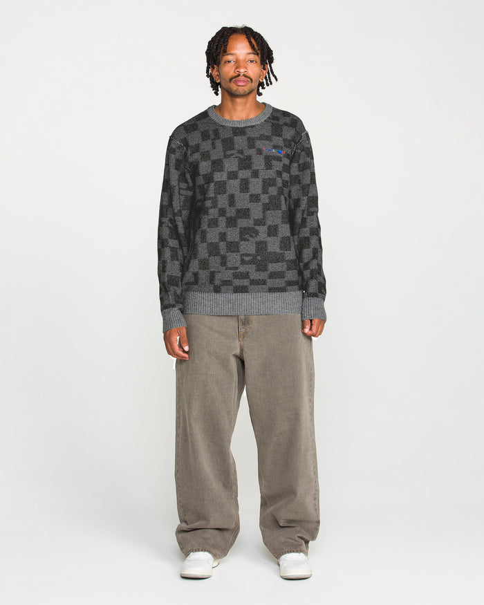 A person stands facing forward against a plain white background, wearing the Volcom Men's Featured Artist Antonio Carrau Sweater with a boxy fit, paired with loose brown pants and white sneakers.