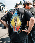 A person outdoors wears the Volcom Men's Featured Artist Daniel Shepard Unlocked Short Sleeve Tee, a black 100% cotton shirt with a colorful hand emerging from barbed wire graphic on the back, standing next to another individual.