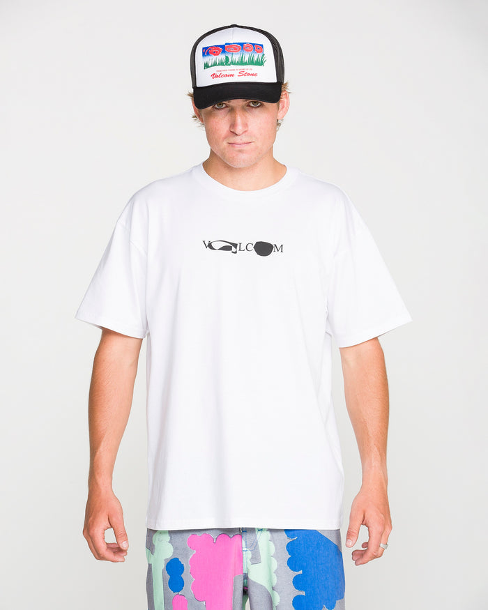 A person wearing a white Volcom Men's Featured Artist Head Check Short Sleeve Tee, crafted from premium ringspun cotton for superior comfort and style and designed by Antonio Carrau, is paired with colorful abstract shorts and a black trucker hat featuring a vibrant graphic design, standing against a plain white background.