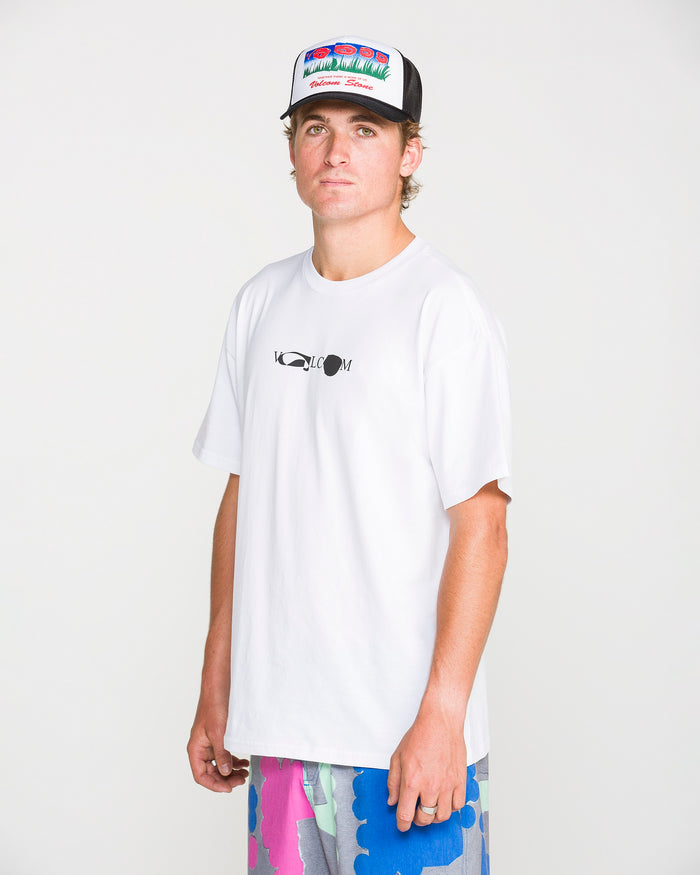 A man wearing a white Volcom Men's Featured Artist Head Check Short Sleeve Tee, crafted from premium ringspun cotton and featuring artwork by Antonio Carrau, stands in front of a plain white background looking at the camera. He pairs the tee with colorful shorts and a multicolored trucker hat.
