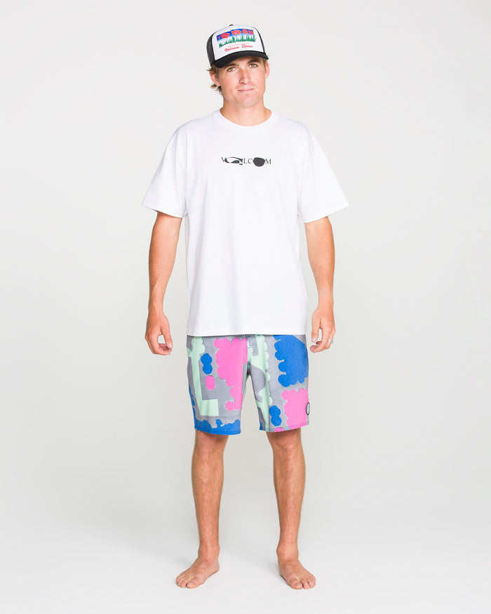 A man stands facing forward against a plain white background, embodying the bold style of Antonio Carrau in a Volcom Men's Featured Artist Head Check Short Sleeve Tee crafted from premium ringspun cotton and adorned with a black logo, paired with colorful pink and blue patterned shorts and a trucker hat.