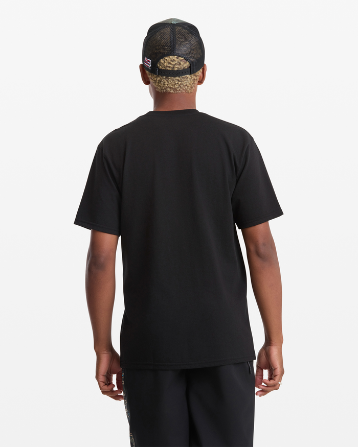 A person stands with their back to the camera, wearing the Volcom Men's Featured Artist Parillo Short Sleeve Tee in black, black shorts, and a black mesh baseball cap with a camouflage-patterned back. The setting is a plain white background.