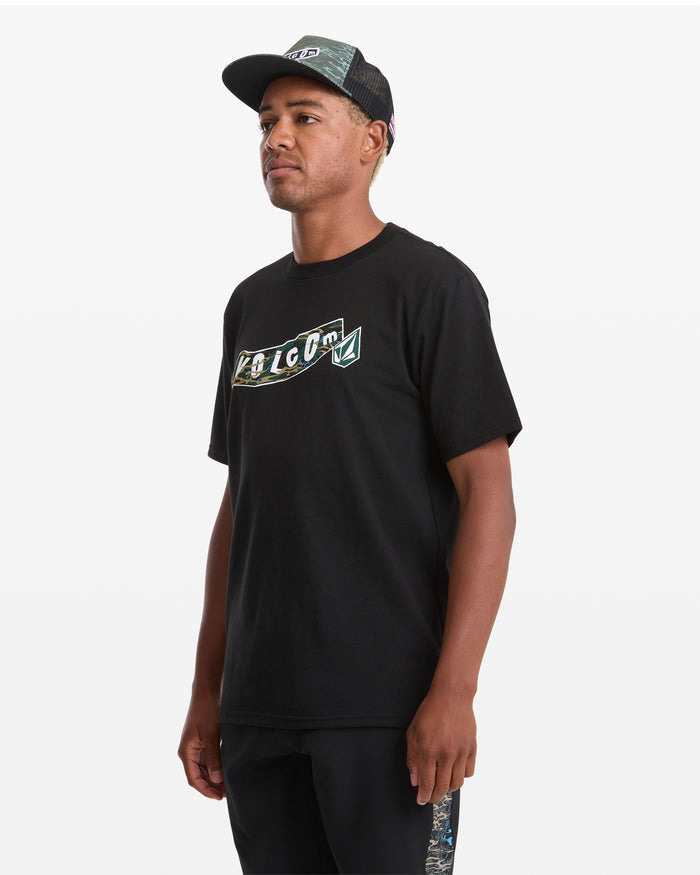 A man wearing a Volcom Men's Featured Artist Parillo Short Sleeve Tee, a black mesh cap, and black shorts stands against a plain white background, looking slightly to his left.