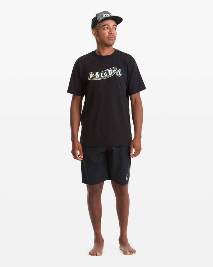 A man stands barefoot against a plain white background wearing a black Volcom Men's Featured Artist Parillo Short Sleeve Tee with a colorful logo, a black cap, and black shorts.