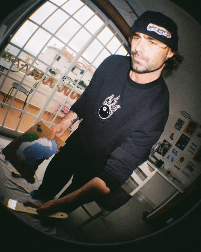 A man in a Volcom Men's Featured Artist Seth Conboy Crew and beanie is painting a canvas indoors, holding a paintbrush and palette by large windows with a balcony railing behind him.