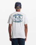 A person wears the Volcom Men's Fed Up Short Sleeve Tee, a classic fit gray 100% cotton shirt with a blue palm tree graphic, "VOLCOM STONE," and "1991" on the back. They pair it with a navy/tan mesh cap and dark shorts.