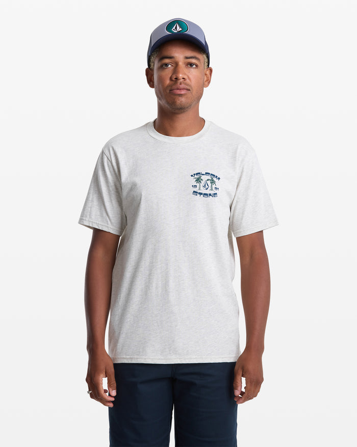 A man stands against a plain white background wearing the Volcom Men's Fed Up Short Sleeve Tee in light gray with a small soft-hand screen print on the left chest, paired with navy shorts and a blue baseball cap.
