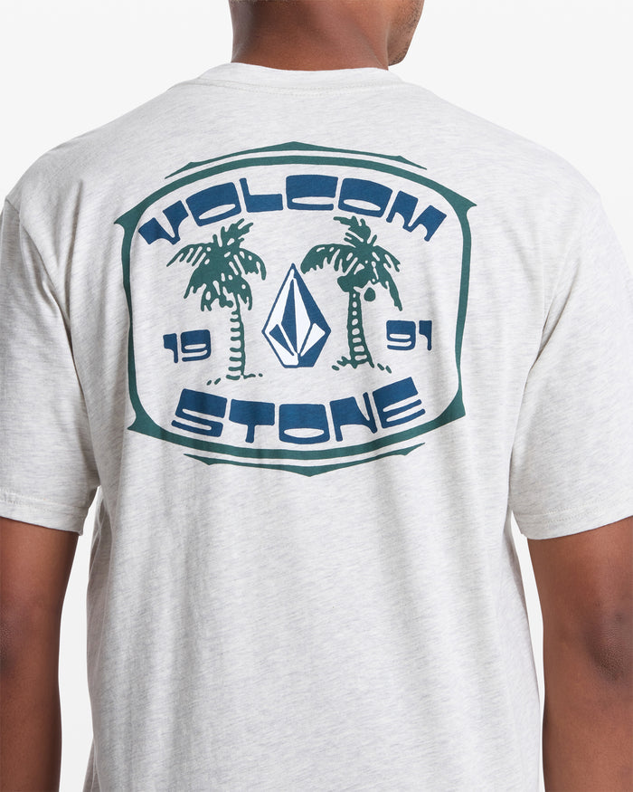 A person wearing the Volcom Men's Fed Up Short Sleeve Tee—a light gray, 100% cotton t-shirt with a soft-hand screen print on the back featuring two palm trees, "VOLCOM STONE," "1991," and a diamond logo. Classic fit for comfort.