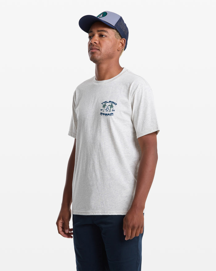 A man wearing the Volcom Men's Fed Up Short Sleeve Tee in light gray with a soft-hand graphic, paired with dark pants and a blue-gray baseball cap, stands against a plain white background.