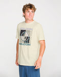 A person wearing the Volcom Men's Filled Out Short Sleeve Tee—a light-colored, modern fit shirt made from soft ring-spun cotton and finished with a bold soft-hand screen-printed graphic logo—pairs it with blue shorts while standing against a plain, light background, gazing off to the side.