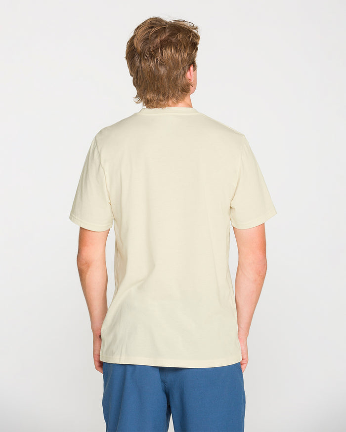 A person with light brown hair is shown from behind, wearing the Volcom Men's Filled Out Short Sleeve Tee in plain off-white and blue shorts, standing against a white background.