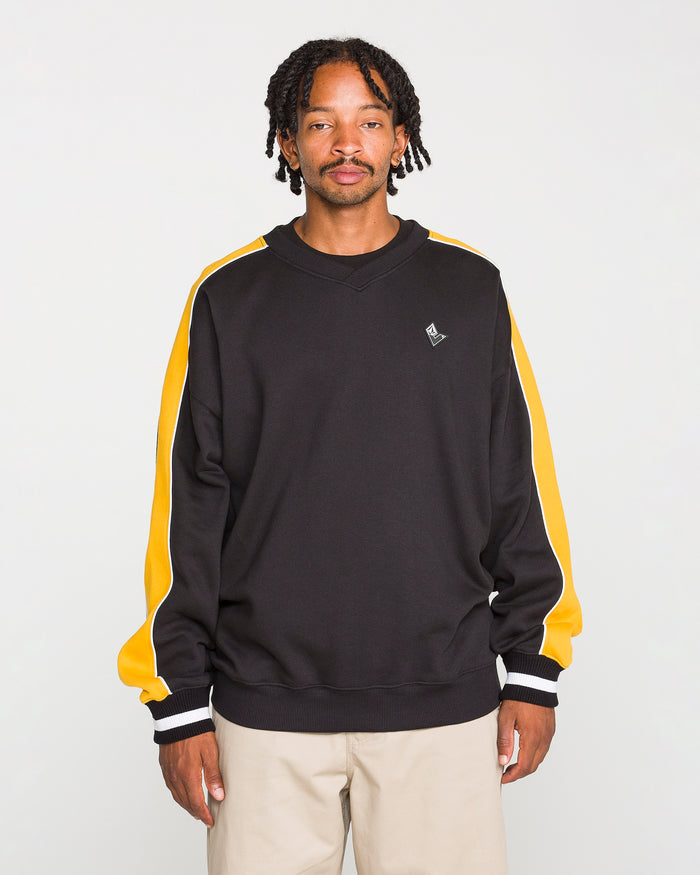 A man with short dreadlocks wears the Volcom Men's Flying Hi Crew, a black sweatshirt with yellow and white colorblocked sleeves crafted from brushed-back fleece with REPREVE recycled polyester, paired with beige pants and standing against a plain light background.