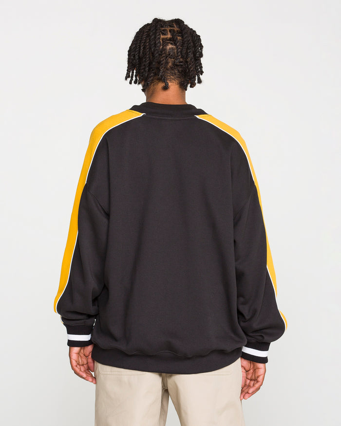 A person is facing away from the camera, wearing the Volcom Men's Flying Hi Crew—a black sweatshirt with colorblocked sleeves featuring yellow and white stripes—paired with light-colored pants, set against a plain white background.
