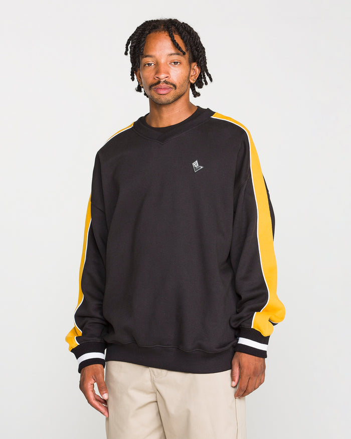 A man with short, twisted hair and a neutral expression stands against a plain background, wearing the Volcom Men's Flying Hi Crew—a black sweatshirt featuring brushed-back fleece and colorblocked sleeves with yellow and white stripes—paired with beige pants. This sweatshirt is crafted from REPREVE recycled polyester for an environmentally conscious touch.