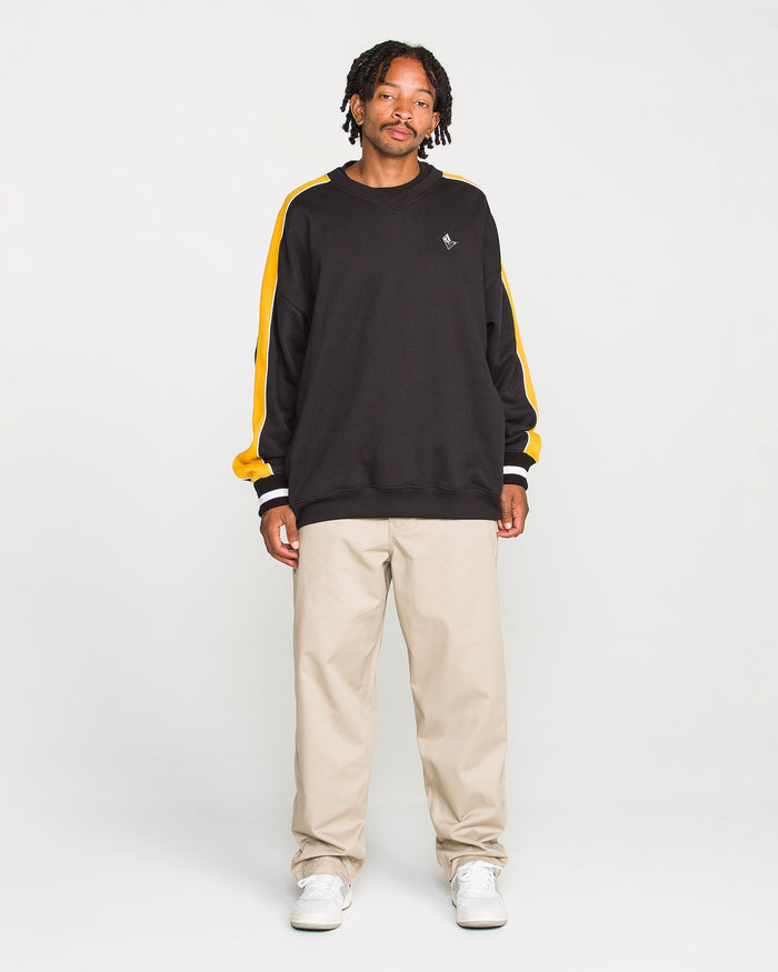 A person stands facing forward, wearing the Volcom Men's Flying Hi Crew—a black sweatshirt with yellow colorblocked sleeves made from brushed-back fleece featuring REPREVE recycled polyester—paired with beige pants and white sneakers against a plain white background.