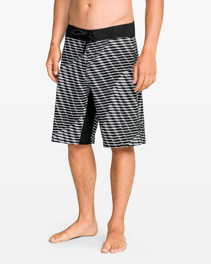 A man stands barefoot against a plain white background wearing Volcom Men's Fomenter Mod board shorts featuring black and white geometric patterns, a black waistband, and 4-way stretch fabric.