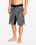 A man stands barefoot against a plain white background wearing Volcom Men's Fomenter Mod board shorts featuring black and white geometric patterns, a black waistband, and 4-way stretch fabric.