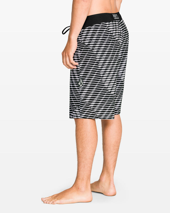 A person stands barefoot in Volcom Men's Fomenter Mod board shorts, black and white geometric pattern with a drawstring waist and left leg logo made from REPREVE recycled fibers, shown from the side and back against a plain background.