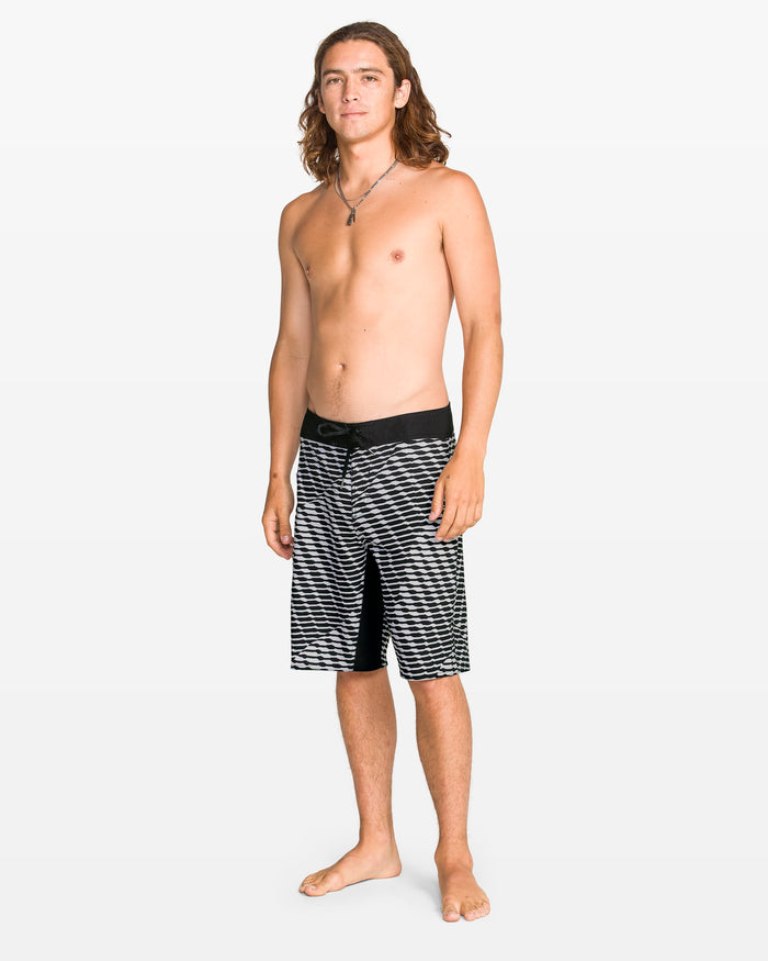 A young man with long brown hair stands barefoot against a plain background, wearing Volcom Men's Fomenter Mod black and white striped board shorts made from REPREVE recycled fibers and a necklace. He faces forward, looking relaxed.