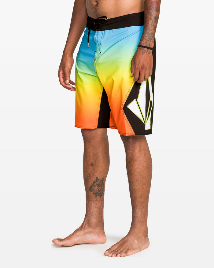 A person stands barefoot wearing Volcom Men's Foster Mod 21" boardshorts with a black waistband and bold geometric leg logo; tattoos are visible on their shin and forearm.